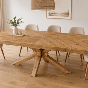 May include: A rectangular light brown wooden dining table with a herringbone pattern on the tabletop and a central cross-legged base, surrounded by six upholstered cream-colored chairs with light brown wooden legs.
