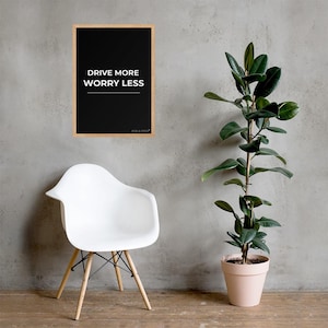 Peut inclure: Une affiche murale rectangulaire dans un cadre en bois clair avec un fond noir et le texte blanc DRIVE MORE WORRY LESS ainsi qu'un petit logo en bas. L'affiche est présentée sur un mur gris au-dessus d'une chaise en plastique blanc.