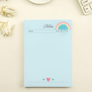 May include: A light blue notepad featuring the word Notes in cursive script at the top, a colorful rainbow and smiling blue cloud graphic, a date line, days of the week initials, and a bottom border with a pink heart flanked by two small blue hearts.