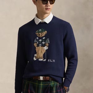 May include: A navy blue long-sleeve jumper featuring a central graphic of a teddy bear wearing a green cap and an argyle vest, with the text RLX embroidered below. The jumper is worn over a white collared shirt, paired with green plaid trousers.