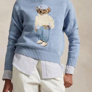 May include: A light blue knit sweater featuring a central graphic of a teddy bear wearing a cap, a cream-colored sweater, and blue jeans. The sweater is layered over a white and blue striped collared shirt, paired with white trousers. Text reads RehneFinds.