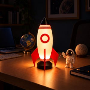 Op de afbeelding: Een raketvormige lamp met een wit lichaam, rode vinnen en een zwarte neuskegel. De lamp geeft een warm licht en heeft een rood cirkelvormig embleem aan de voorzijde. Daarnaast staat een klein wit astronautenfiguurtje.