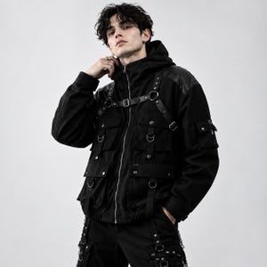 K&ouml;nnte beinhalten: Eine schwarze Kapuzenjacke mit Frontrei&szlig;verschluss, mehreren Utility-Taschen mit Druckkn&ouml;pfen und einem integrierten Gurtsystem mit verstellbaren Riemen, Metallschnallen und D-Ringen &uuml;ber Brust und Schultern.