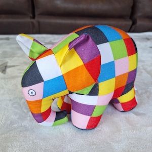 May include: A plush elephant toy featuring a vibrant patchwork design of multicolored squares in shades of red, orange, yellow, green, blue, purple, pink, white, and black. The elephant has large ears, a trunk, and a single visible eye with a black pupil.