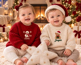May include: A red knit jumper with a snowman and the name Leni, and a cream knit jumper with a teddy bear and the name Jasper. Both jumpers feature embroidered holiday designs and are worn by two children in a festive setting.
