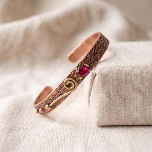 May include: A copper cuff bracelet featuring a hammered, textured surface, a single round faceted deep red gemstone, and a decorative brass wire spiral accent with small brass spheres at the end of the wire detail on the metal band.