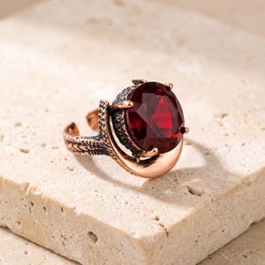 Puede incluir: Un anillo con una gran piedra preciosa de color rojo intenso y corte ovalado, engastada en una montura de metal oscuro y texturizado. El aro tiene un acabado pulido y liso en la parte inferior y un patrón grabado oscuro alrededor de la piedra.