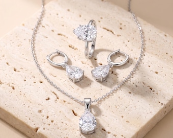 Op de afbeelding: Een zilverkleurige sieradenset bestaande uit een hanger met een peervormige heldere steen aan een fijne ketting, een bijpassende ring met peervormige steen en twee oorbellen met scharniersluiting, voorzien van briljante witte stenen.