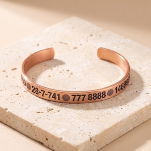 May include: A copper-toned metal cuff bracelet featuring an open design. The exterior surface is engraved with black text and symbols, specifically the sequence 28-7-741, circular icons, the number 777, the number 8888, and the number 148999.