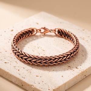 Può includere: Un bracciale color rame caratterizzato da un design a catena intrecciata con finitura metallica. Il bracciale è composto da maglie intrecciate che creano un aspetto strutturato simile a una corda ed è fissato con una chiusura a moschettone.