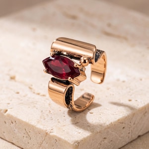 May include: A gold-toned open ring featuring a marquise-cut deep red gemstone set horizontally. The band has a smooth, polished surface on the outer edges and a dark, textured, hammered finish on the inner sides, creating a bold, structural design.