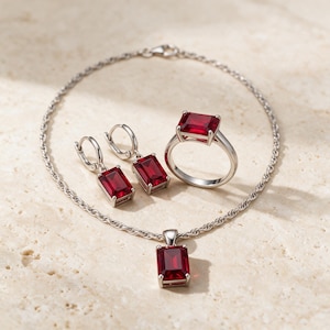 May include: A jewelry set featuring a silver-toned rope chain necklace with a rectangular red gemstone pendant, a matching ring with a rectangular red gemstone, and two drop earrings with rectangular red gemstones in silver settings.