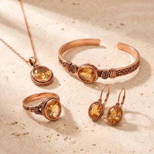 May include: A jewellery set in rose-toned metal featuring a necklace with an oval pendant, an open cuff bracelet, a ring, and a pair of lever-back earrings. Each piece is set with a faceted oval citrine gemstone and displays intricate metalwork.