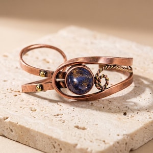 May include: A copper cuff bracelet featuring a circular blue and gold speckled stone centerpiece. The band is constructed from two overlapping copper strips with small brass rivets and a decorative twisted wire accent near the stone setting.