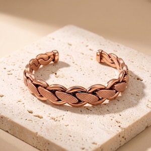 May include: A rose gold-toned cuff bracelet featuring a braided design with alternating smooth and cross-hatched textured links. The bracelet has an open-ended construction, allowing for adjustment, and displays a metallic finish throughout.