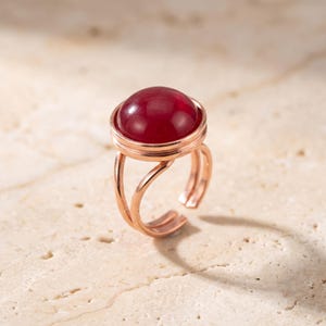 Peut inclure: Une bague couleur or rose ornée d'une pierre cabochon ronde, lisse et rouge foncé, sertie dans une monture circulaire. L'anneau est composé de deux fils métalliques fins et parallèles qui se courbent pour former une ouverture réglable.