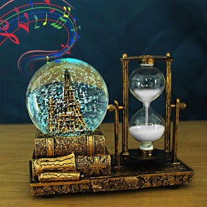 May include: A decorative desk ornament featuring a glass snow globe with a miniature Eiffel Tower inside, resting on a stack of textured bronze-colored books next to a rotating hourglass filled with white sand, all set on a matching base.