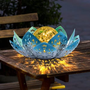 May include: A lotus-shaped solar light featuring blue metal petals with intricate cut-out patterns surrounding a central crackle glass globe. The globe contains warm yellow LED string lights that cast a glowing reflection onto the surface below.