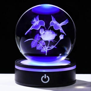 May include: A clear glass sphere featuring a 3D laser-etched design of two hummingbirds hovering over a rose with leaves and buds. The sphere rests on a black circular base with a glowing blue light and a white power button icon on the front.