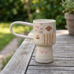 May include: A ceramic mug with a tall, flared base and a large, rounded handle. The exterior features a patchwork design with hand-painted illustrations of a small brown bird, a cupcake with a red cherry, pink spots, and tan checkered squares.