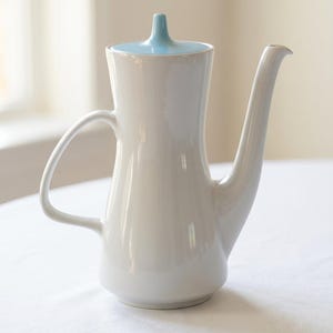 Op de afbeelding: Een witte keramische koffiepot met een hoog, slank lichaam, een gebogen handvat en een lange, taps toelopende tuit. Het deksel heeft een contrasterende lichtblauwe kleur en is voorzien van een kleine, puntige knop in het midden.