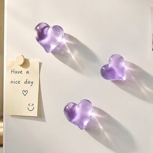 May include: Three translucent purple heart-shaped magnets attached to a white surface. A rectangular cream-colored note is held by a circular magnet, featuring the handwritten text "Have a nice day" with a heart and a smiley face drawing.