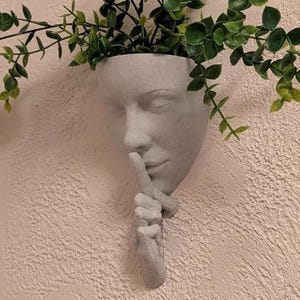 May include: A light gray wall-mounted planter shaped like a human face with a hand held up to the lips in a shushing gesture. The planter has a speckled texture and contains a green leafy plant extending from the top opening of the head.