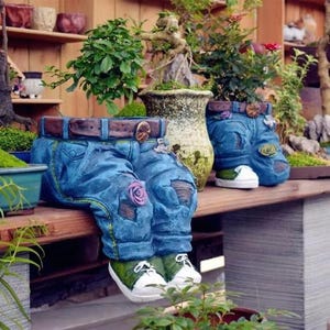 Puede incluir: Una maceta con forma de pantalón vaquero azul que presenta un cinturón marrón con hebilla, detalles desgastados en las rodillas con pequeños adornos de flores moradas y zapatillas verdes con cordones y suelas blancas para colocar en bordes.