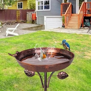 May include: A copper-coloured metal bird bath featuring a round basin with a decorative bird figure on the rim and leaf accents on the stand. A small bird is perched on the edge while another bird is splashing in the water inside the basin.