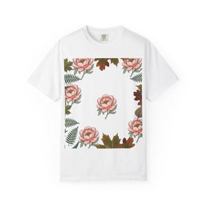 Könnte beinhalten: Ein weißes kurzärmeliges T-Shirt mit einem zentralen Grafikdruck aus rosa Pfingstrosen, umgeben von einem Rand aus grünen Farnwedeln und braunen Herbstblättern. Das Design ist in einem quadratischen Rahmen mit einer einzelnen Pfingstrose angeordnet.