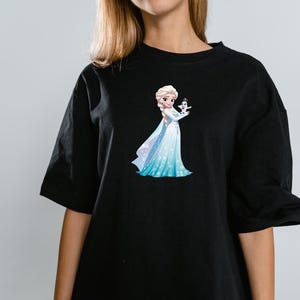 Peut inclure: Un t-shirt noir &agrave; manches courtes avec une impression graphique repr&eacute;sentant un personnage blond en robe bleu clair et blanche tenant une petite figurine de bonhomme de neige blanc. Le personnage a de longs cheveux tress&eacute;s et une robe &agrave; motifs flocons.
