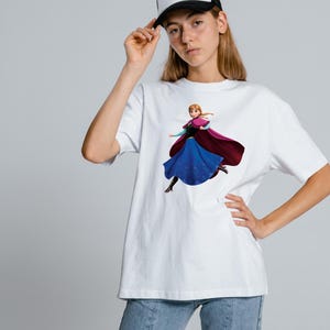 Peut inclure: Un t-shirt blanc &agrave; manches courtes avec une impression graphique repr&eacute;sentant un personnage aux cheveux tress&eacute;s, portant une cape magenta fonc&eacute;, un corsage noir et une longue jupe bleue. Le personnage est dans une pose dynamique.