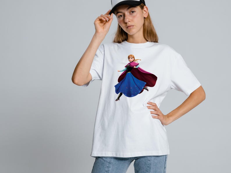 Peut inclure: Un t-shirt blanc &agrave; manches courtes avec une impression graphique repr&eacute;sentant un personnage aux cheveux tress&eacute;s, portant une cape magenta fonc&eacute;, un corsage noir et une longue jupe bleue. Le personnage est dans une pose dynamique.