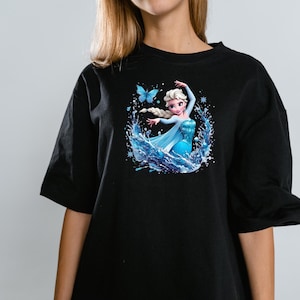 Peut inclure: Un t-shirt noir &agrave; manches courtes avec une impression graphique repr&eacute;sentant un personnage blond avec une tresse, portant une robe bleue, entour&eacute; d'&eacute;claboussures d'eau, de flocons de neige et d'un papillon bleu centr&eacute; sur la poitrine.