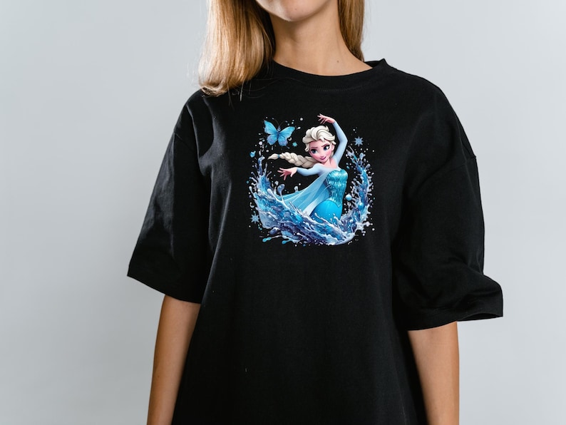 Peut inclure: Un t-shirt noir &agrave; manches courtes avec une impression graphique repr&eacute;sentant un personnage blond avec une tresse, portant une robe bleue, entour&eacute; d'&eacute;claboussures d'eau, de flocons de neige et d'un papillon bleu centr&eacute; sur la poitrine.
