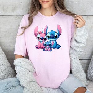 May include: A light pink short-sleeved t-shirt featuring a graphic print of three blue and pink alien characters. The central design depicts two larger characters sitting beside a smaller baby character, all with large eyes and prominent ears.