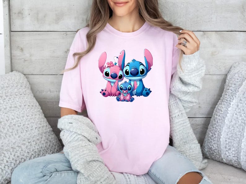 May include: A light pink short-sleeved t-shirt featuring a graphic print of three blue and pink alien characters. The central design depicts two larger characters sitting beside a smaller baby character, all with large eyes and prominent ears.
