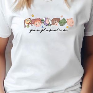 May include: A white short-sleeved t-shirt featuring a graphic print of six cartoon character heads arranged in a row above the black cursive text "you've got a friend in me" and small colorful stars scattered around the character illustrations.