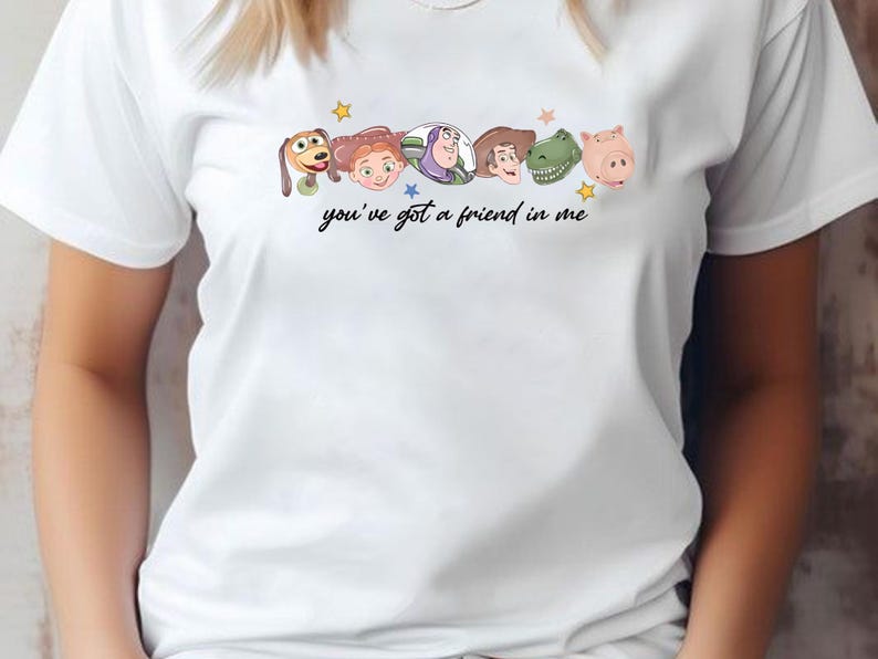 May include: A white short-sleeved t-shirt featuring a graphic print of six cartoon character heads arranged in a row above the black cursive text "you've got a friend in me" and small colorful stars scattered around the character illustrations.