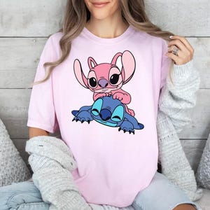 May include: A light pink short-sleeved t-shirt featuring a graphic print of a blue alien character lying down with a smaller pink alien character sitting on its back. The design is centred on the chest of the shirt, showcasing both characters.