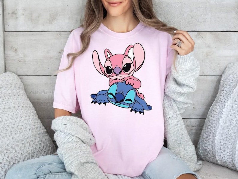 May include: A light pink short-sleeved t-shirt featuring a graphic print of a blue alien character lying down with a smaller pink alien character sitting on its back. The design is centred on the chest of the shirt, showcasing both characters.