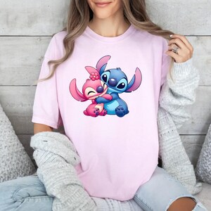 May include: A light pink short-sleeved t-shirt featuring a graphic print of two cartoon characters, one blue and one pink, embracing each other. The pink character has a small pink flower on its head, and both characters have large ears.