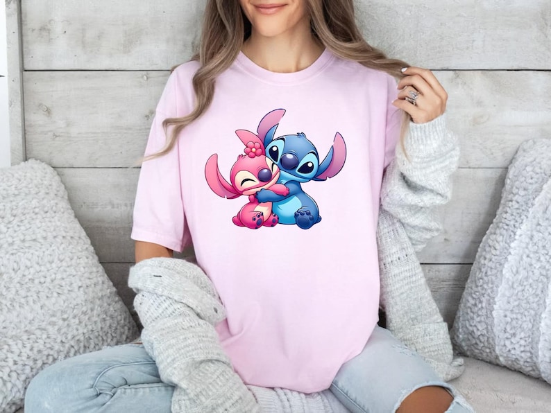 May include: A light pink short-sleeved t-shirt featuring a graphic print of two cartoon characters, one blue and one pink, embracing each other. The pink character has a small pink flower on its head, and both characters have large ears.