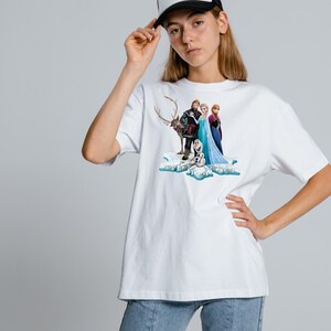 Peut inclure: Un t-shirt blanc &agrave; manches courtes avec une impression graphique des personnages Elsa, Anna, Kristoff, Sven et Olaf de Frozen debout sur une formation de glace bleue et blanche. Le t-shirt a un col rond et une coupe d&eacute;contract&eacute;e.