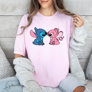 May include: A light pink short-sleeved t-shirt featuring a graphic print of two cartoon characters, one blue and one pink, facing each other. The characters have large ears and are depicted in a seated position on the centre of the chest area.