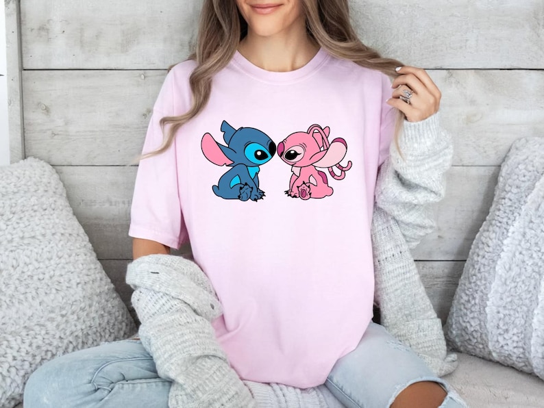 May include: A light pink short-sleeved t-shirt featuring a graphic print of two cartoon characters, one blue and one pink, facing each other. The characters have large ears and are depicted in a seated position on the centre of the chest area.