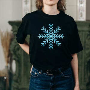 Peut inclure: Un t-shirt noir &agrave; manches courtes arborant un grand motif central de flocon de neige bleu clair avec un aspect cristallin textur&eacute;. Le t-shirt pr&eacute;sente une encolure ronde classique et est port&eacute; avec un jean bleu fonc&eacute; et une ceinture noire.