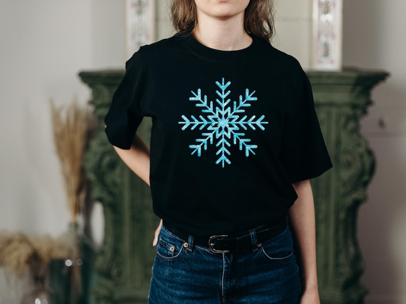 Peut inclure: Un t-shirt noir &agrave; manches courtes arborant un grand motif central de flocon de neige bleu clair avec un aspect cristallin textur&eacute;. Le t-shirt pr&eacute;sente une encolure ronde classique et est port&eacute; avec un jean bleu fonc&eacute; et une ceinture noire.