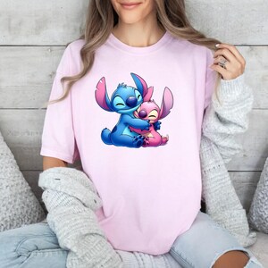 May include: A light pink short-sleeved t-shirt featuring a graphic print of the blue character Stitch hugging the pink character Angel. The design is centred on the chest of the shirt, showcasing both characters with closed eyes and smiling expressions.