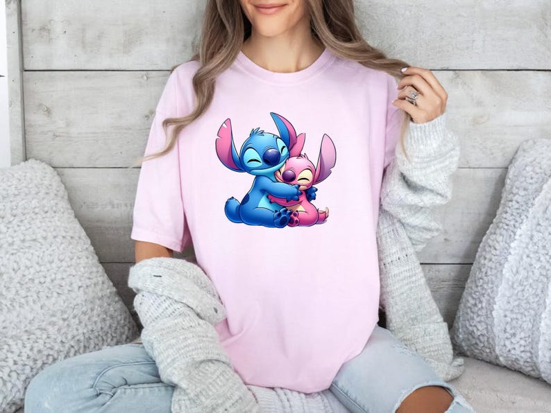 May include: A light pink short-sleeved t-shirt featuring a graphic print of the blue character Stitch hugging the pink character Angel. The design is centred on the chest of the shirt, showcasing both characters with closed eyes and smiling expressions.
